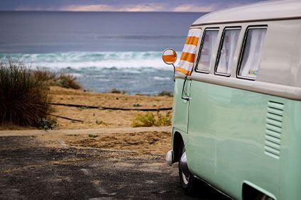 En minibuss i mintgrönt med vitt tak, står på en parkeringsplats som vetter mot en strand och hav med små vågor som slår mot stranden. I horisonten syns en mörkgrå himmel.