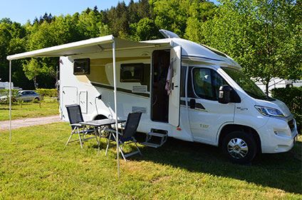 En vit husbil står parkerad på gräs med gröna träd i bakgrunden. Husbilen står med öppen sidodörr och ett separat extra tak över ett set med campingstolar och bord i svart.