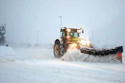 En gul traktor plogar snö på en väg medan snö virvlar runt. Himlen är gråmulen och gatubelysningen är tänd.