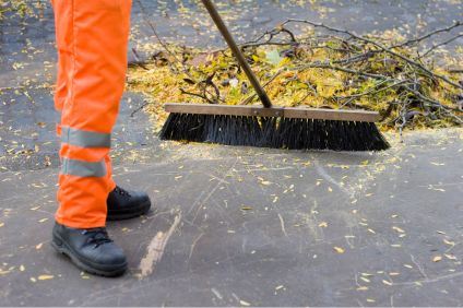 En person sopar upp löv och kvistar. Personen har orange varselbyxor.