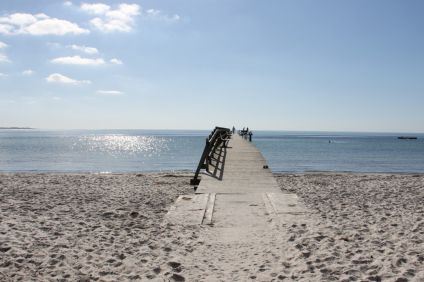 Lång träbrygga som går ut från en sandstrand mot ett hav där solen lyser genom några disiga moln.