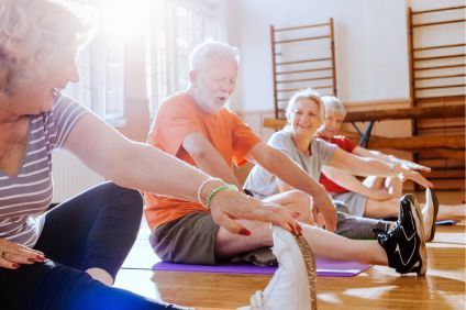 Äldre kvinnor och män sitter med benen utsträckta och stretchar. De är i en gymnastiksal med trägolv och solen strålar in från fönster högt upp.