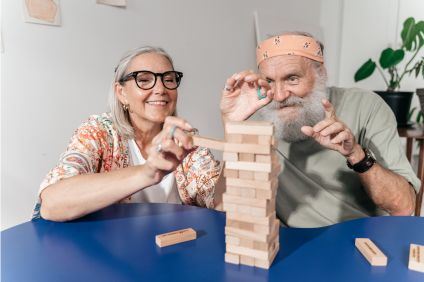 En gråhårig kvinna och en man i grått långt hår och bandana spelar Jenga, ett spel där man staplar trästavar till ett torn. De har roligt ihop.