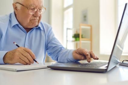 En senior man i ljusblå skjorta sitter med en laptop och skriver i ett block.