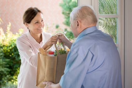 En kvinna i brunt hår ler medan hon sträcker över en matkasse till en  senior man.