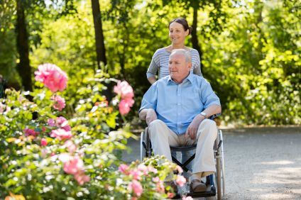 En senior man i blå skjorta och grå byxor sitter i en rullstol. Han skjutsas runt bland stora blommor av en brunhårig kvinna i grårandig tröja.