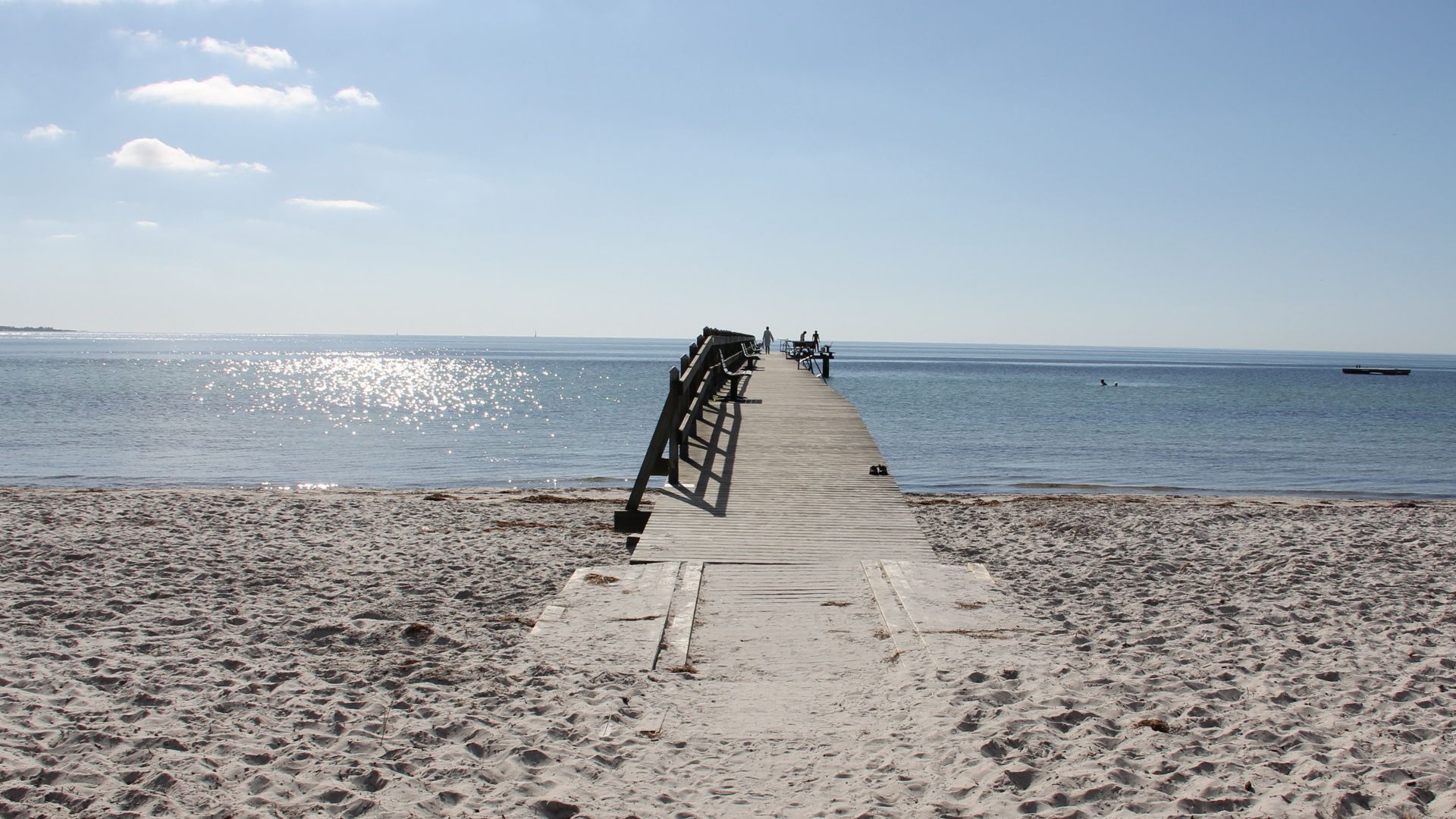 Träbrygga som leder ut från sandstrand långt ut i havet. På bryggan syns några människor längst bort. Solen skiner och några vita moln syns på den blå himlen.