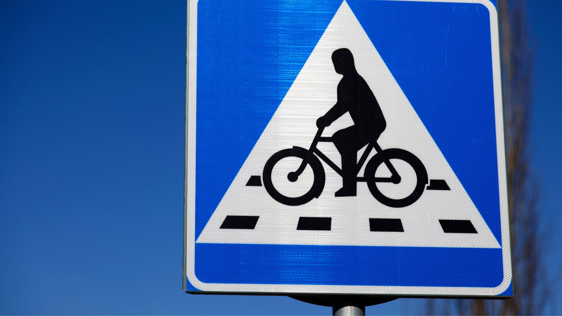 Skylt för cykelöverfart där en svart figur sitter på en cykel och far över streckad gata. Bakom skylten är det en blå himmel.