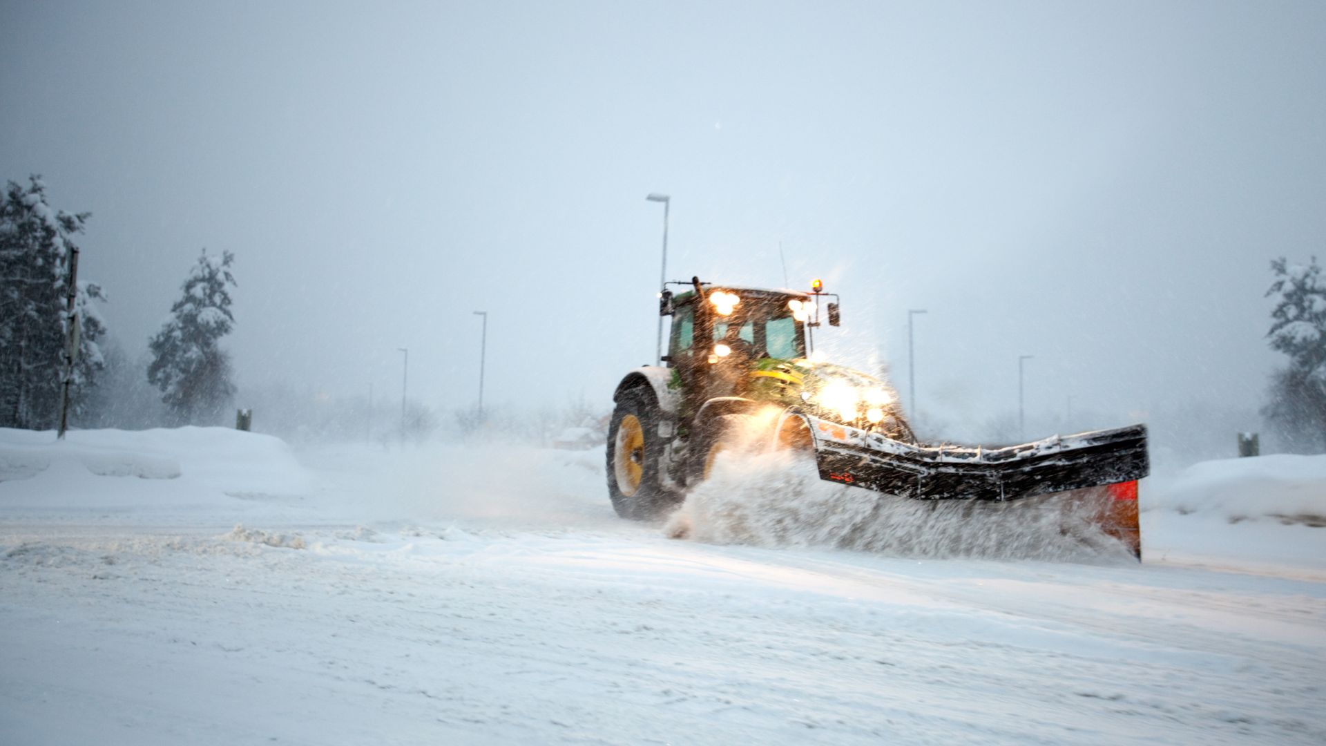 En gul traktor plogar snö på en väg medan snö virvlar runt. Himlen är gråmulen och gatubelysningen är tänd.