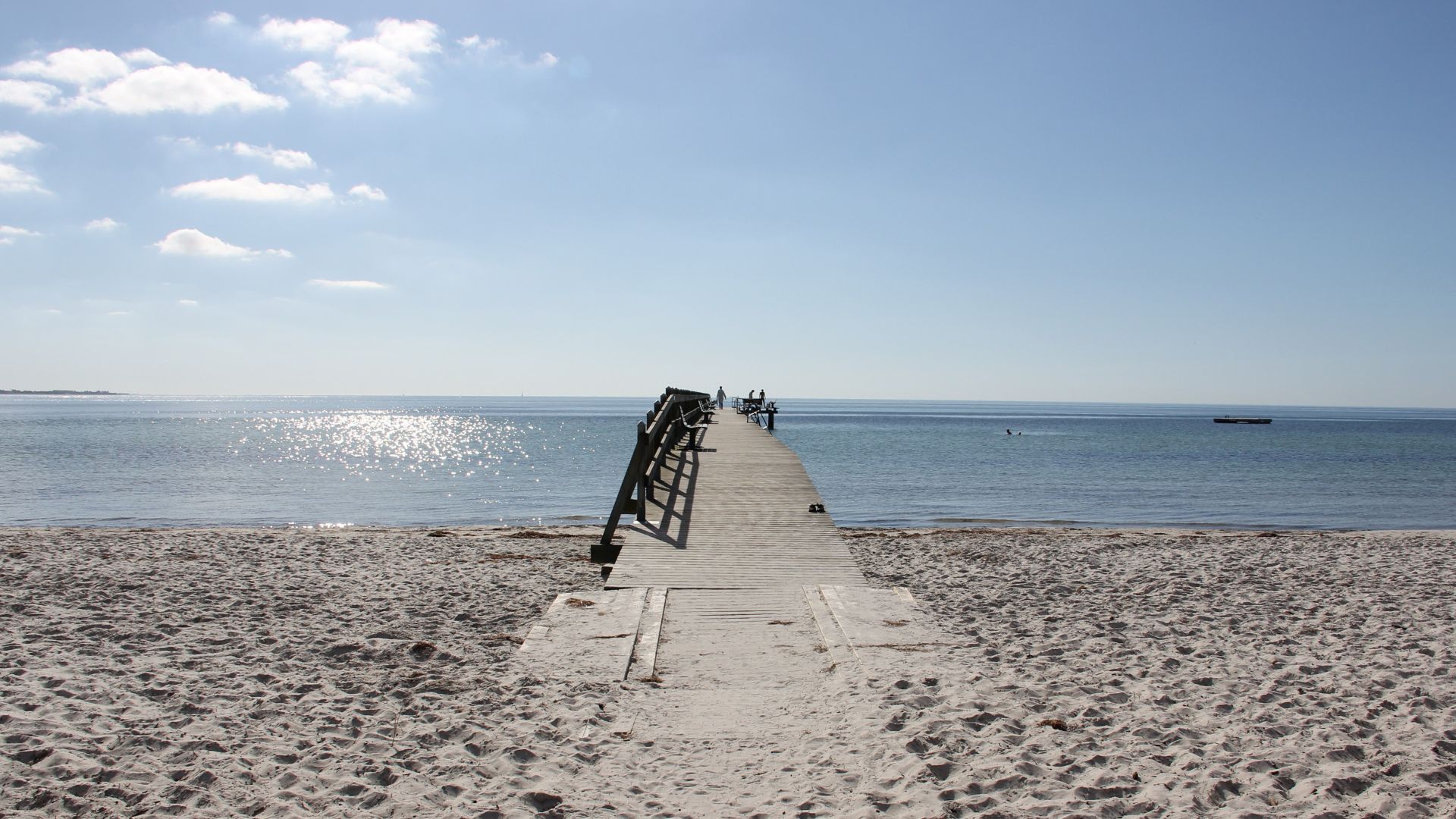 Träbrygga leder från en bred sandstrand ut i vattnet. Det är en solig dag.